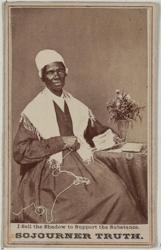 Sojourner Truth seated beside a table with a vase of flowers. She is looking at the camera and holding yarn in one hand.