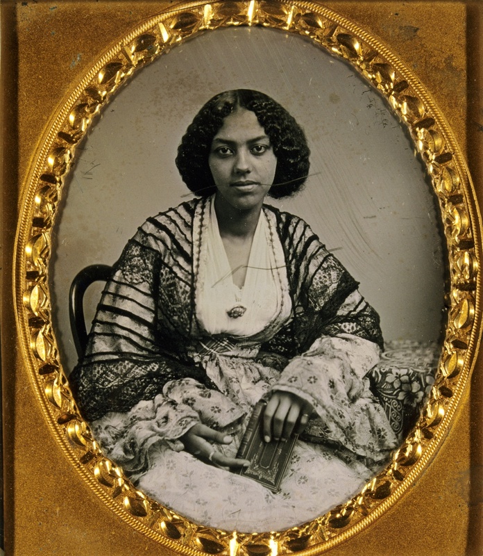 3/4 body photo of a seated African American woman. She is wearing elaborate attire and holding a book. The portrait is in a gold frame.