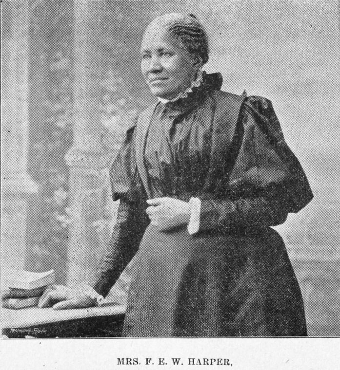 1/2 body photo of Frances Ellen Watkins Harper standing and looking left, her left hand on a table and her right hand at her waist.