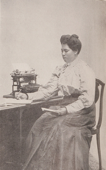 3/4 body photo of L. A. J. Moorer seated, holding a pen in one hand and a book in the other.