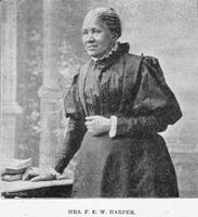 1/2 body photo of Frances Ellen Watkins Harper standing and looking left, her left hand on a table and her right hand at her waist.
