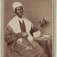 Sojourner Truth seated beside a table with a vase of flowers. She is looking at the camera and holding yarn in one hand.