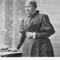 1/2 body photo of Frances Ellen Watkins Harper standing and looking left, her left hand on a table and her right hand at her waist.