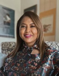 A Black woman with light brown skin and long brown hair wearing a dark floral blouse.